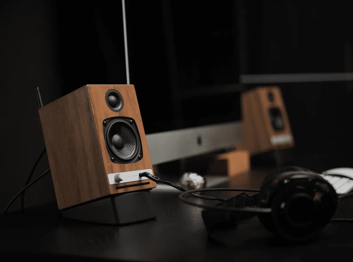 Walnut Audioengine speakers on a dark desk beside a monitor and headphones